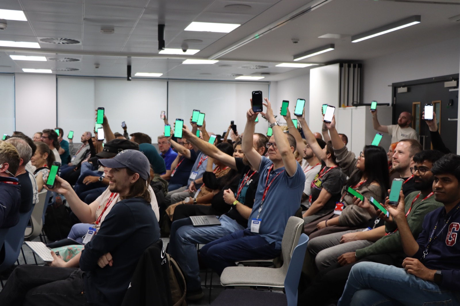 A room of people holding their phones up, screens lit green in unison, during a pixelmesh effect